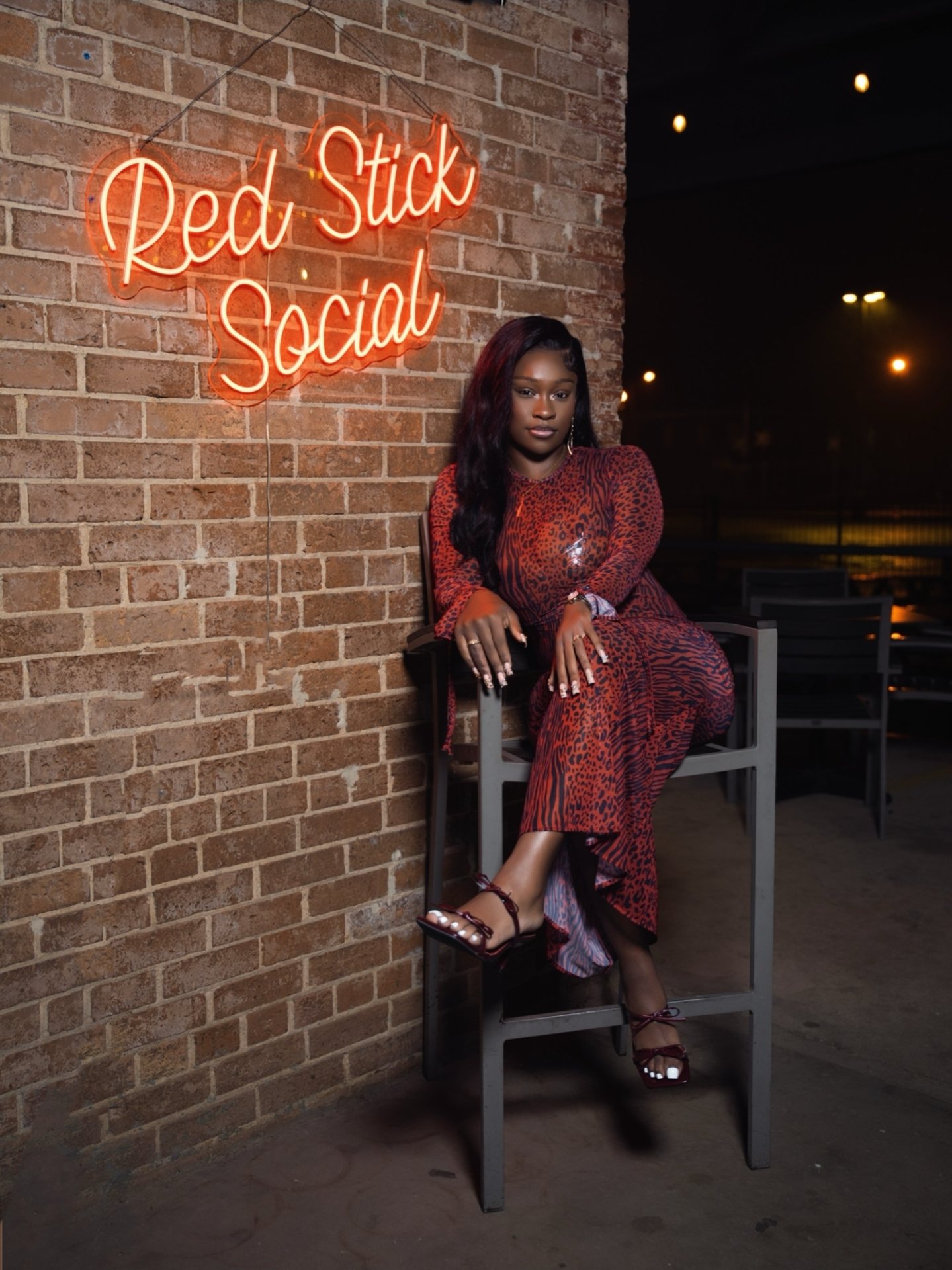 Dyimond seated on a tall chair under the Red Stick Social neon sign