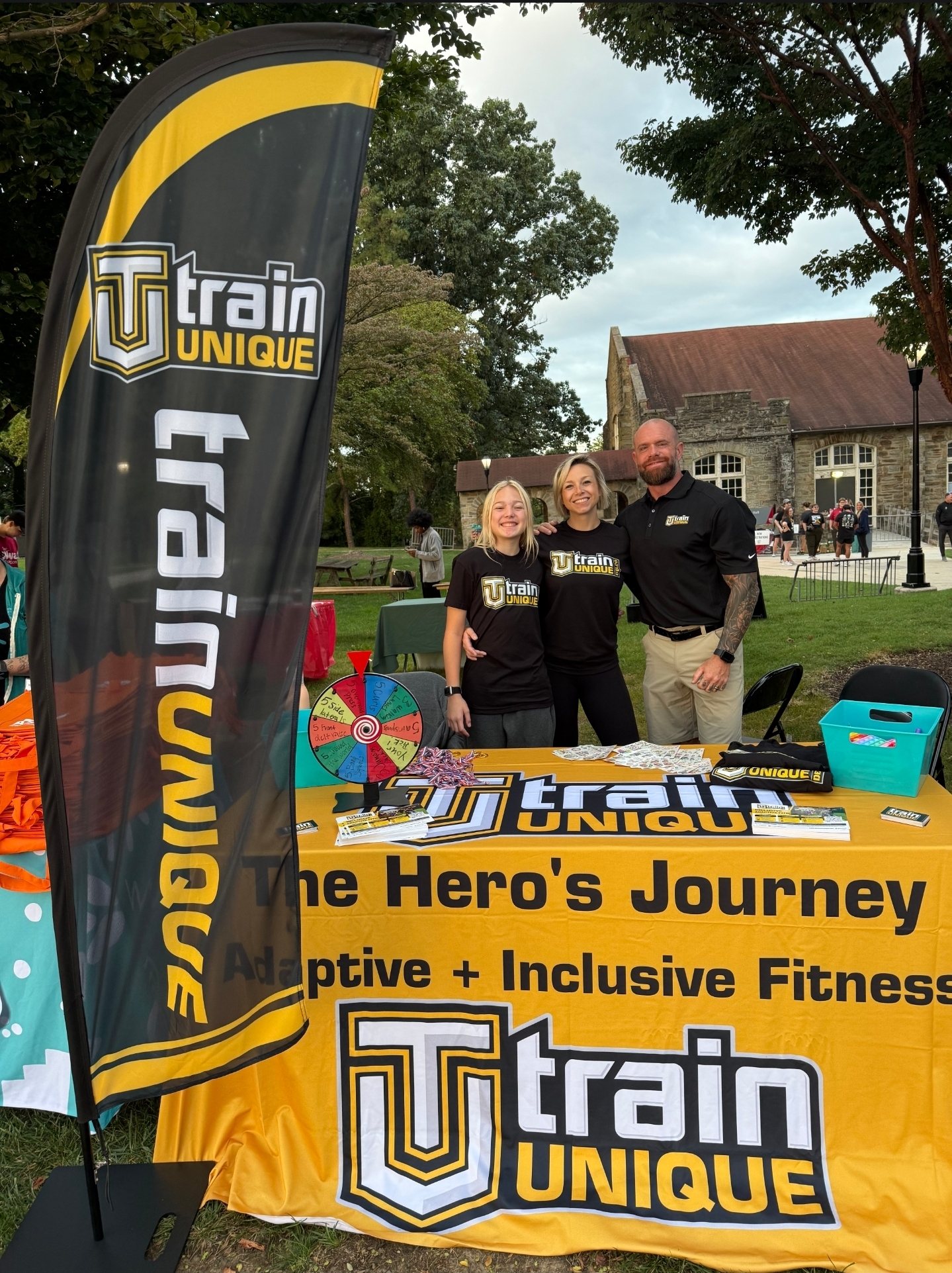 Kevin at the Train Unique table at an outdoor community event, banner and spin wheel set up