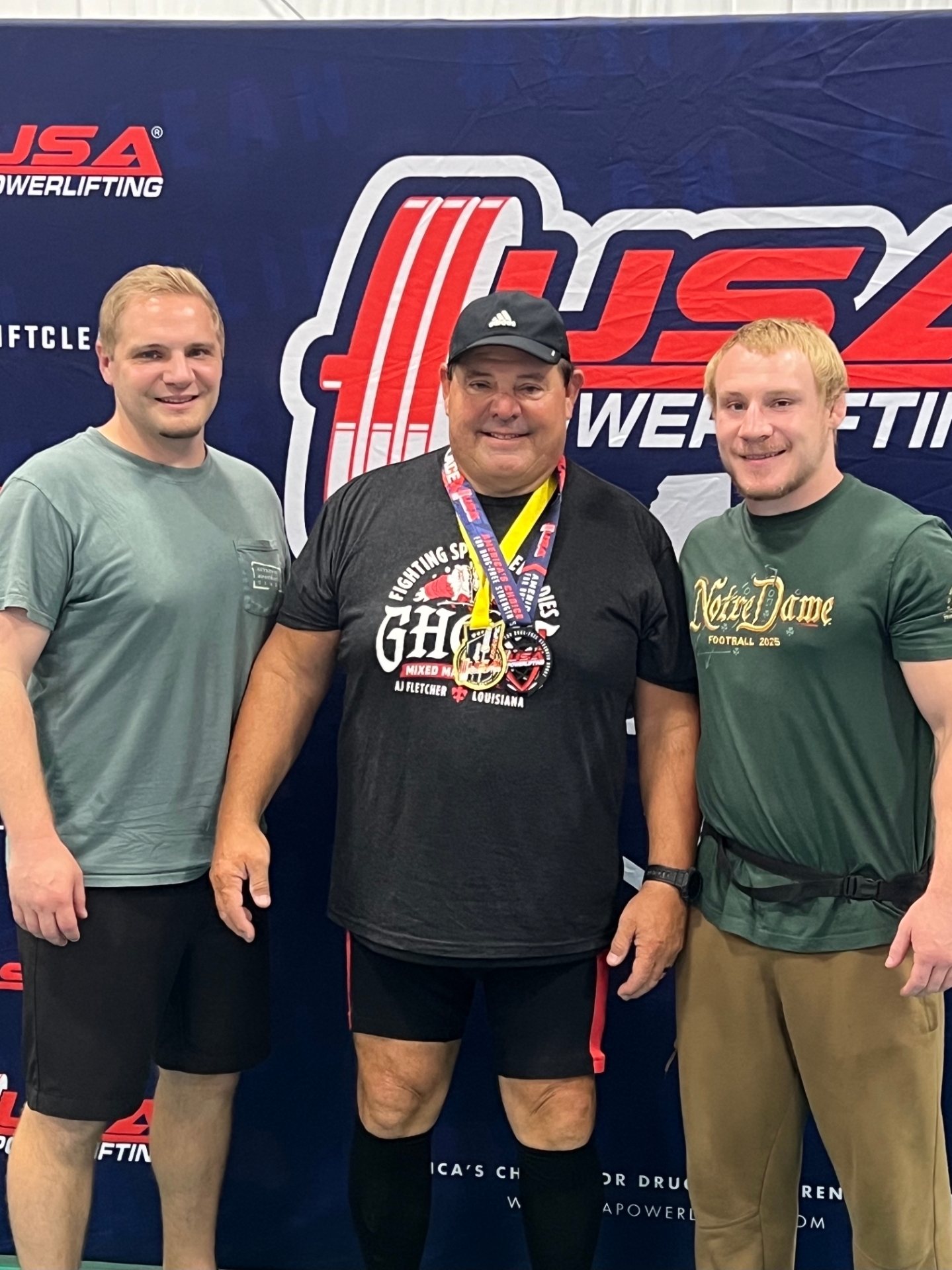 Paul with two medals around his neck at a USA Powerlifting banner, two young lifters beside him
