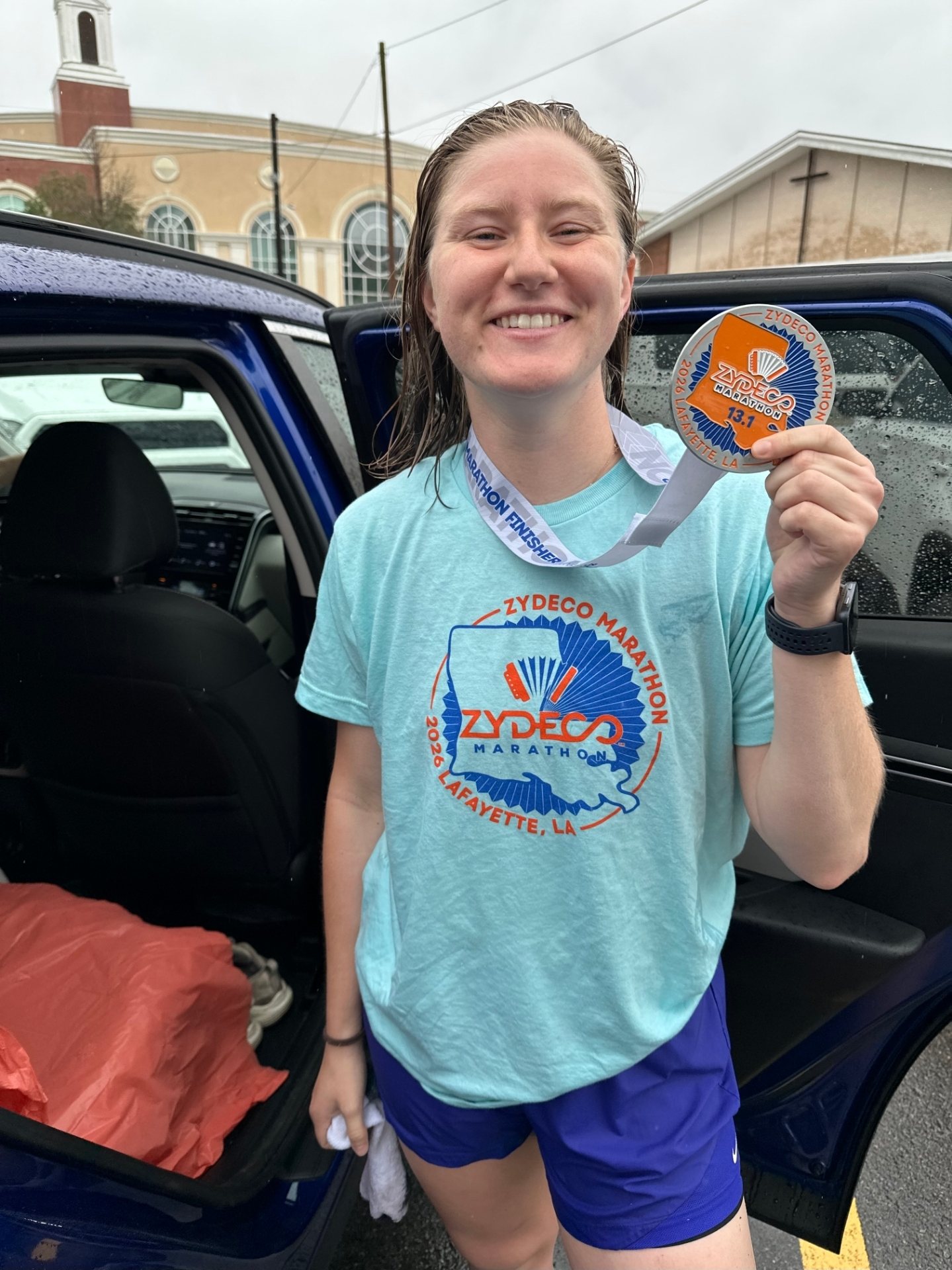 Coach Shannon, soaked and smiling, holding up her Zydeco Marathon half-marathon medal in Lafayette